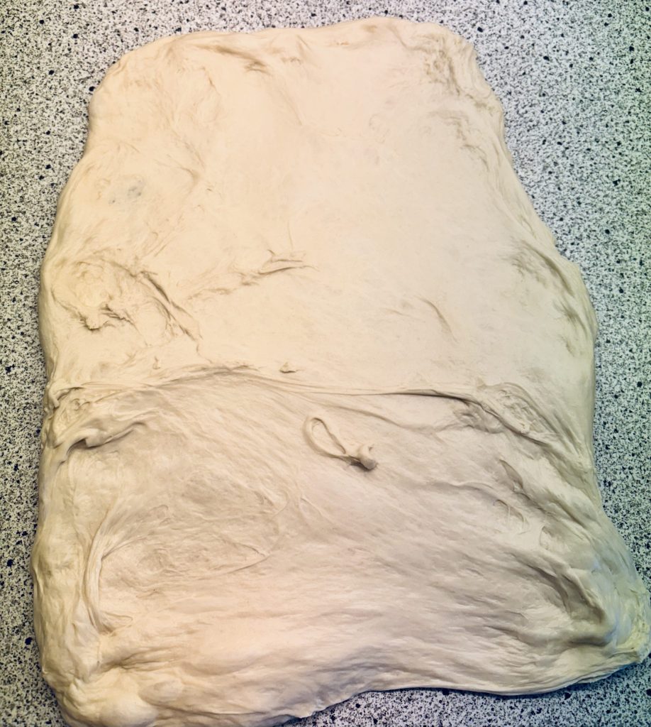 Eine große, flache Teigfläche aus Sauerteig, die auf einer Arbeitsplatte aus grauem Material ausgebreitet ist.