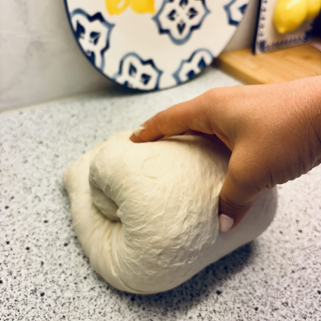 Eine Hand, die einen gut durchgekneteten Teig auf einer Arbeitsfläche mit einer strukturierten Oberfläche hält, im Hintergrund ein farbenfrohes Tellerdesign.