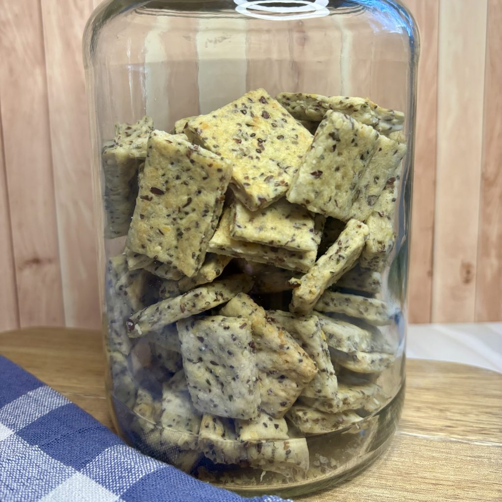 Ein Glas mit knusprigen Sauerteigcrackern auf einem Holztisch, mit einem karierten Tuch daneben.