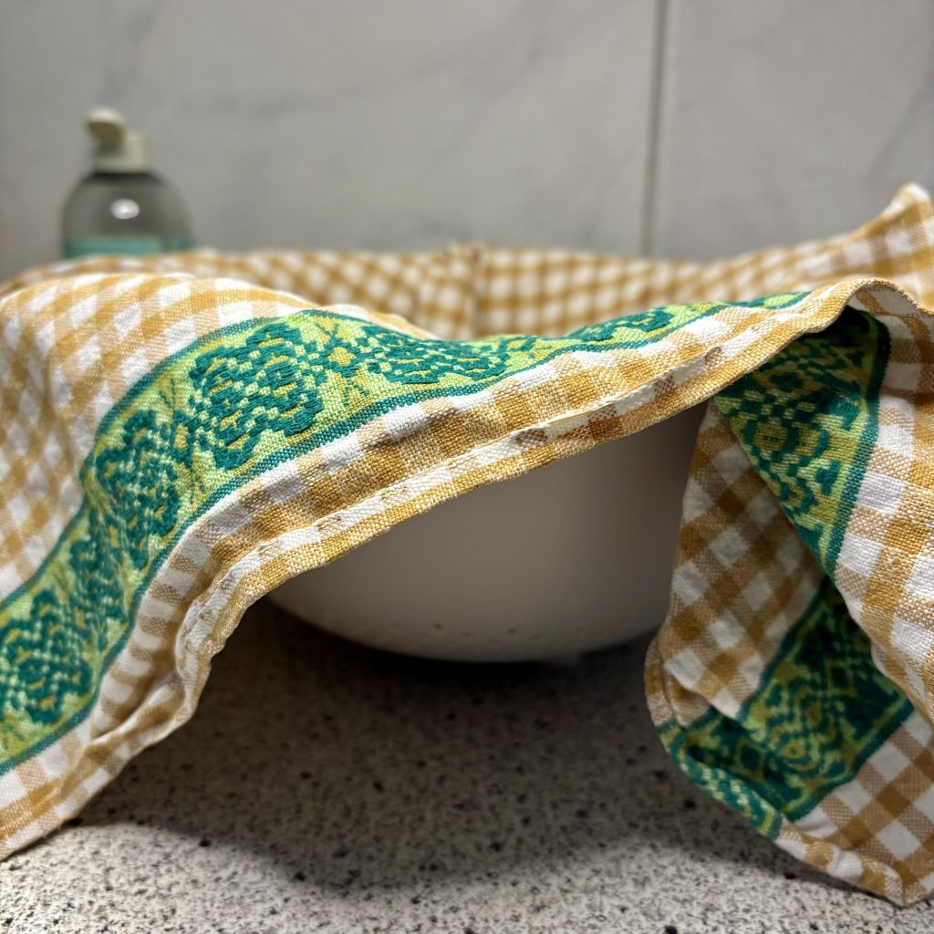 A close-up of a bowl covered with a patterned kitchen towel, resting on a granite countertop. A bottle of liquid soap is visible in the background.