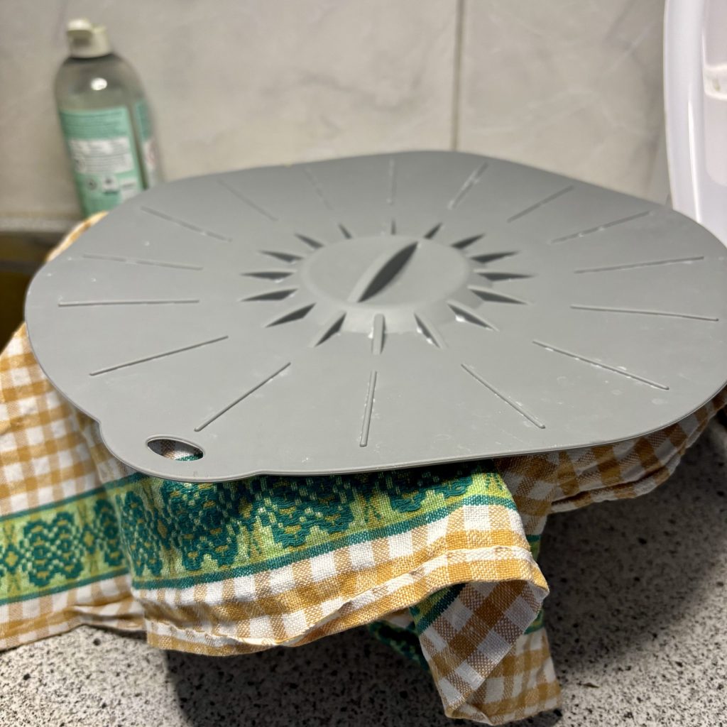 A gray silicone lid with a sunburst pattern placed on a checkered cloth, sitting on a kitchen counter next to a bottle of cleaning solution.