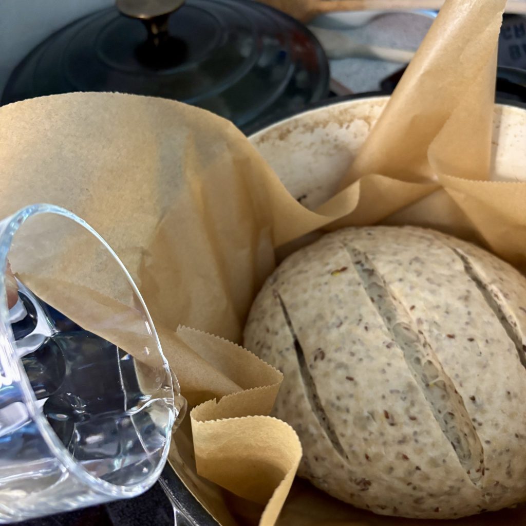A round loaf of bread dough in a pot lined with parchment paper, with shallow cuts on the surface, and a glass of water positioned nearby.