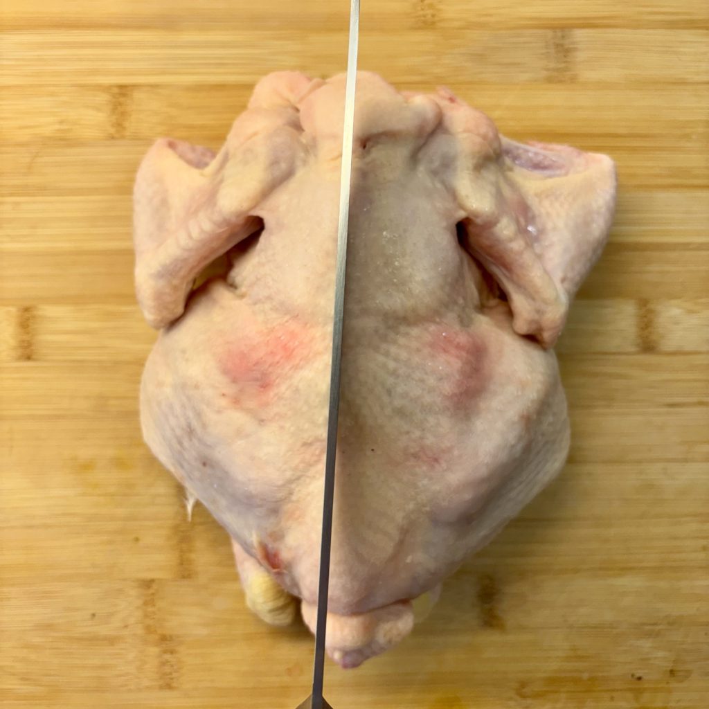 Ein frisches Hähnchen, das auf einem Holzbrett liegt, mit einem scharfen Messer, das vertikal in der Mitte angesetzt ist.