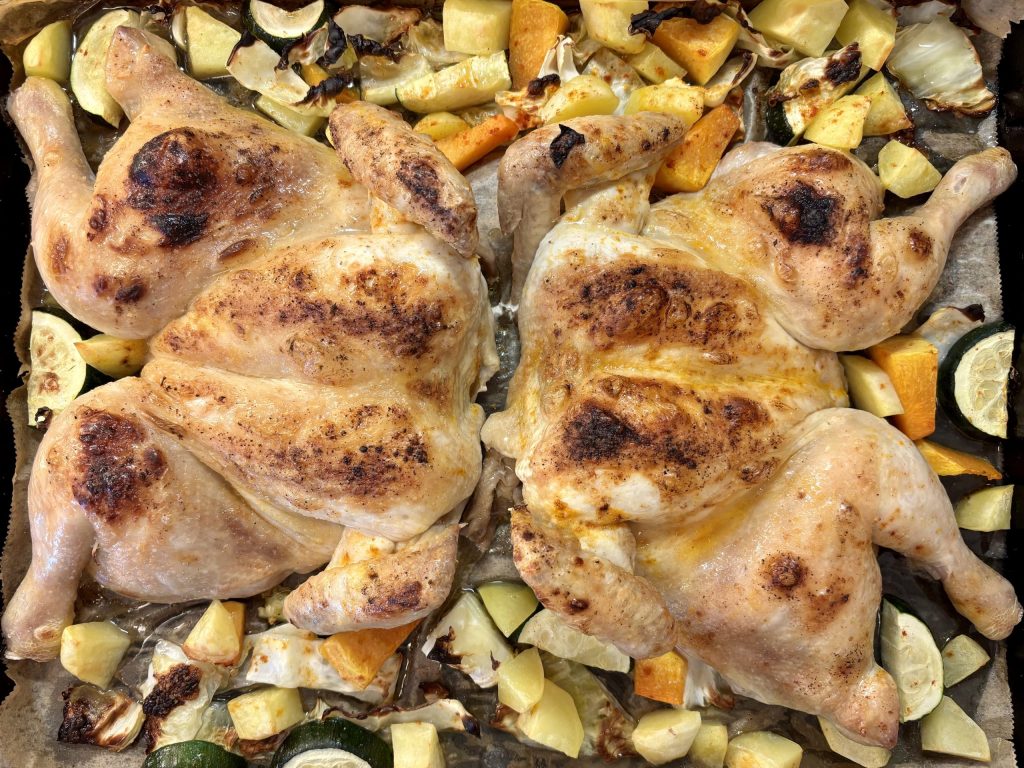 Zwei gegrillte Hähnchen auf einem Blech, umgeben von verschiedenen Gemüsesorten wie Kürbis, Zucchini und Kartoffeln.
