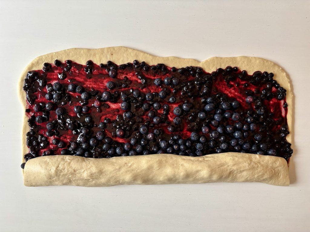 Ein ausgerollter Teig mit einer Schicht aus Heidelbeermarmelade und frischen Heidelbeeren, auf einer hellen Oberfläche.
