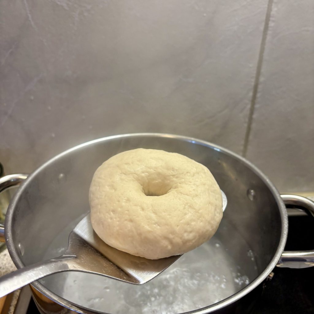Ein ungebackenes Bagel wird mit einem Schaumlöffel aus einem Topf mit kochendem Wasser gehoben.
