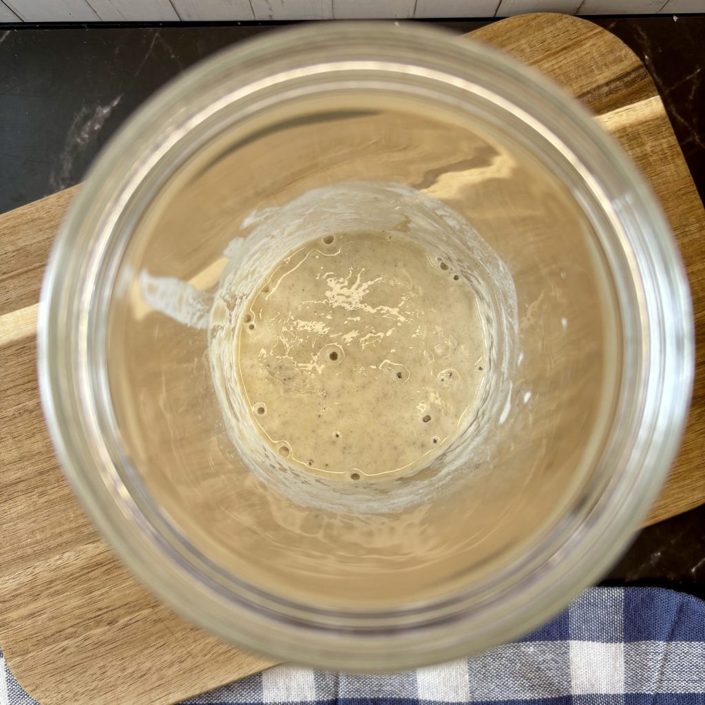 Obere Ansicht eines Glasgefäßes mit fermentierendem Sauerteig, das auf einem Holzbrett steht.